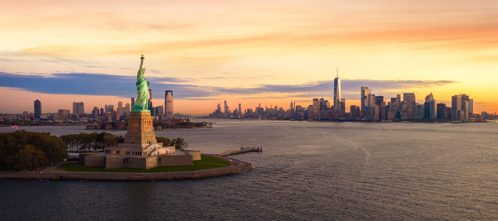 The Statue of Liberty on its island at sunset, with the Lower Manhattan skyline across the harbor
