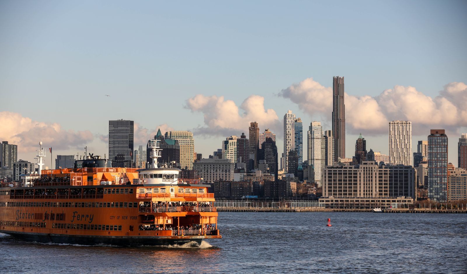 An orange Staten Island Ferry full of passengers crossing New York Harbor, with the Brooklyn waterfront and skyline behind
